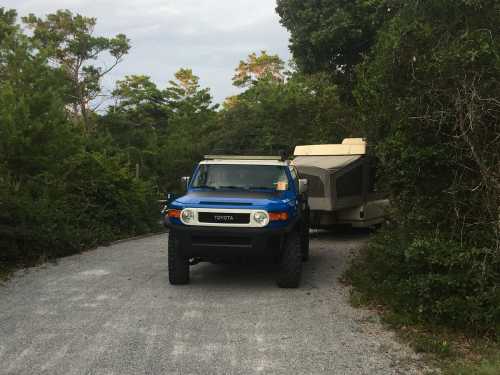 A blue Toyota FJ Cruiser parked on a gravel road, towing a small camper trailer surrounded by greenery.