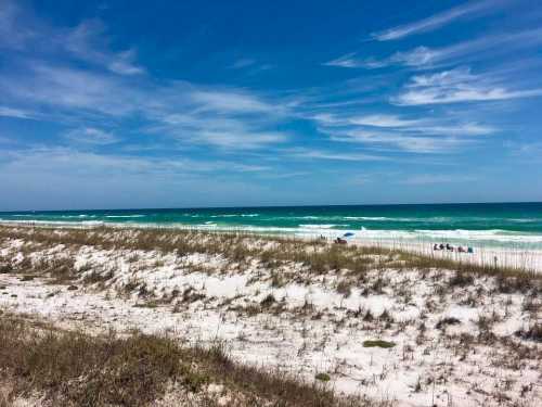 A serene beach scene with white sand, gentle waves, and a clear blue sky, dotted with a few beachgoers in the distance.