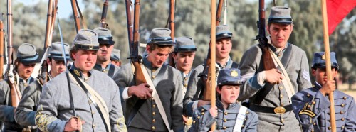 A group of young men in Civil War-era uniforms, holding rifles and a red flag, marching together.