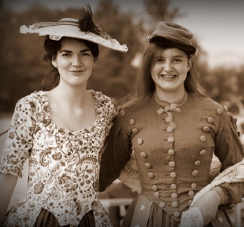 Two women in vintage dresses, smiling and standing together outdoors, captured in a sepia-toned photograph.