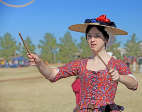A young woman in a historical dress and hat holds two sticks, performing outdoors with a clear blue sky in the background.