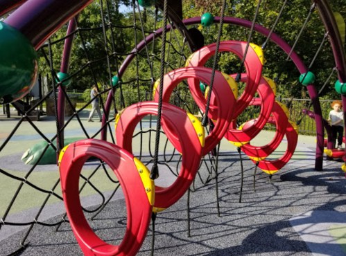 Colorful playground equipment featuring red rings and climbing ropes against a green park backdrop.