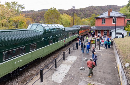 A vintage train at a station with passengers boarding, surrounded by trees and hills in the background.