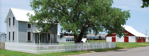 A gray house with a white fence and a large red barn nearby, surrounded by green grass and trees under a clear sky.