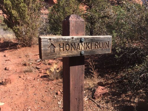 Wooden signpost reading "Honanki Ruin" in a desert landscape with shrubs and red rock formations.