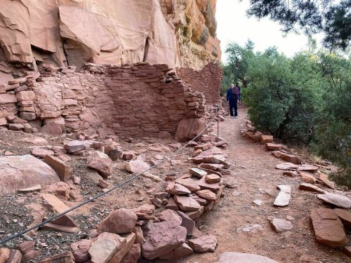Ancient stone ruins along a dirt path, surrounded by greenery and rocky terrain. Two people are visible in the background.