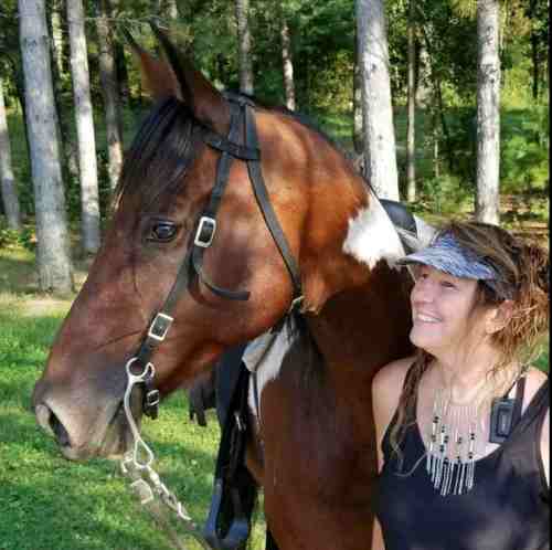 A woman stands beside a brown and white horse in a forested area, smiling and wearing a hat and jewelry.