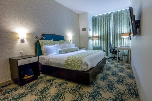 A modern hotel room featuring a queen bed, desk, and large windows with patterned curtains.
