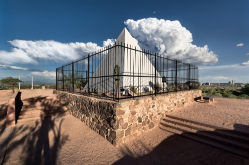 A white pyramid structure surrounded by a black fence, set against a blue sky with clouds and desert landscape.