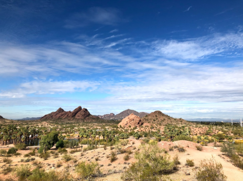 A desert landscape featuring rocky hills, sparse vegetation, and a blue sky with scattered clouds.