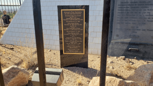 A plaque commemorating Arizona's first governor, surrounded by rocks and a fence, with a pyramid structure in the background.