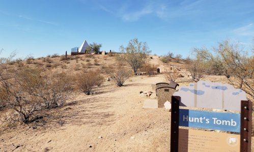 A desert landscape featuring Hunt's Tomb sign, with a small hill and a white structure in the background. Sparse vegetation surrounds.