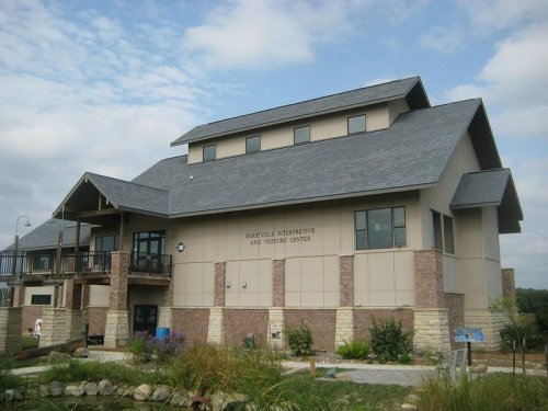 A modern building with a sloped roof, featuring a stone base and large windows, surrounded by greenery and a pond.
