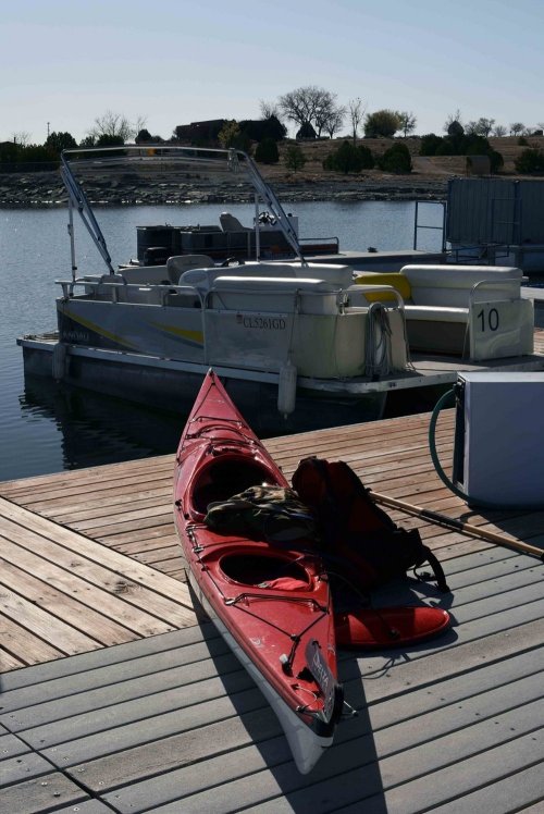 A red kayak rests on a wooden dock beside a pontoon boat on a calm lake. Trees line the shore in the background.