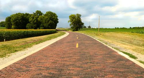 A winding brick road surrounded by green fields and trees under a cloudy sky.