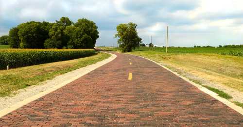 A winding brick road surrounded by fields and trees under a cloudy sky.