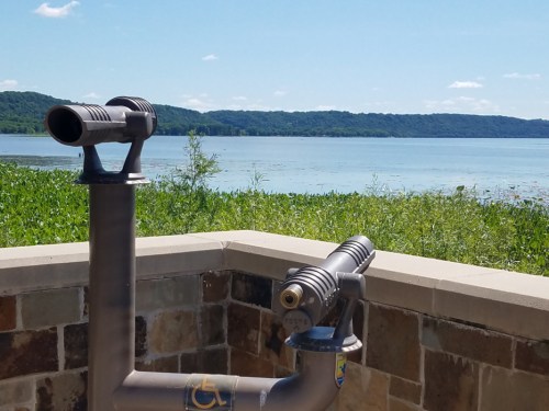 A scenic view of a lake with a telescope mounted on a stone railing, surrounded by greenery and hills in the background.