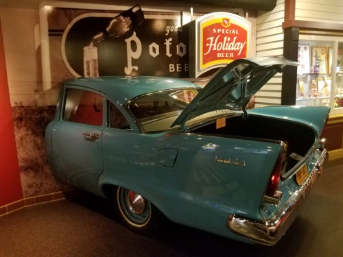 A vintage blue car with its trunk open, displayed in a retro-themed setting with beer advertisements in the background.