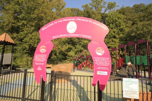 Sign at the entrance of the Medieval Dragon Playground, South Bowie Community Center, surrounded by playground equipment.