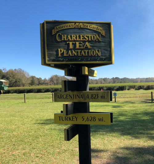 Sign for Charleston Tea Plantation, listing distances to Argentina (4,823 mi) and Turkey (5,628 mi). Green landscape in background.