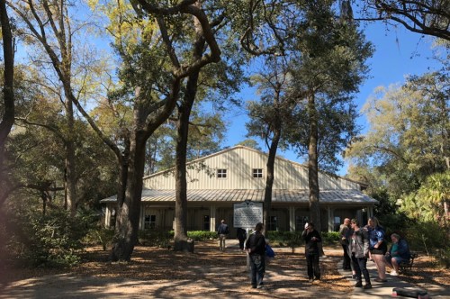 A group of people walks near a large, light-colored building surrounded by trees on a sunny day.