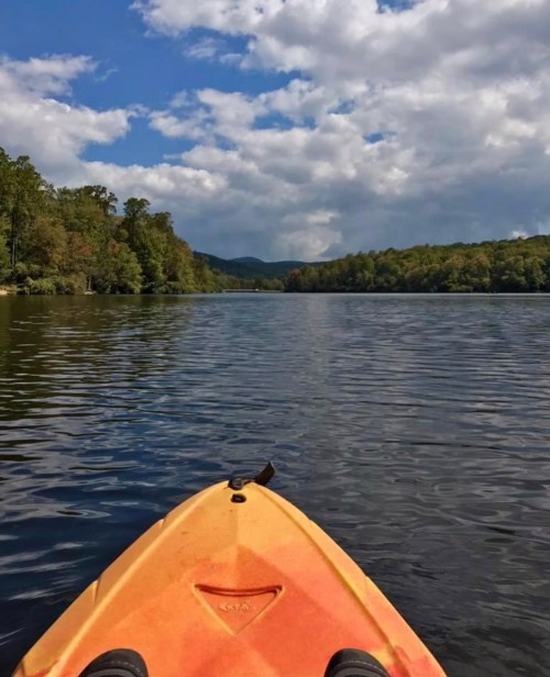A kayak on calm water, surrounded by trees and mountains under a partly cloudy sky.