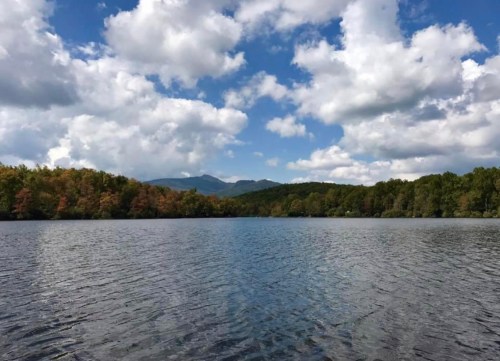 A serene lake surrounded by trees with autumn colors, under a blue sky filled with fluffy white clouds.