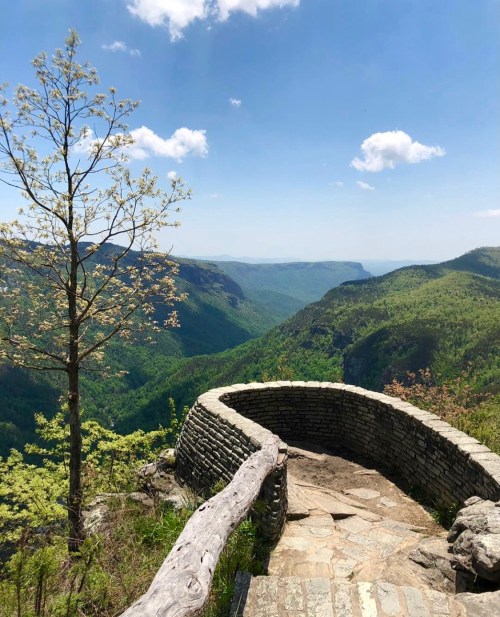 A scenic overlook with a stone railing, surrounded by lush green mountains and a clear blue sky.