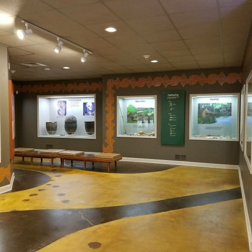 Interior of a museum exhibit featuring display cases and benches, with informational panels on gathering and hunting.