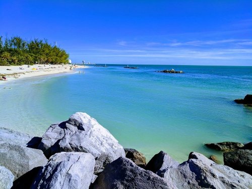 A serene beach scene with clear blue water, rocky foreground, and a sunny sky, dotted with people enjoying the shore.