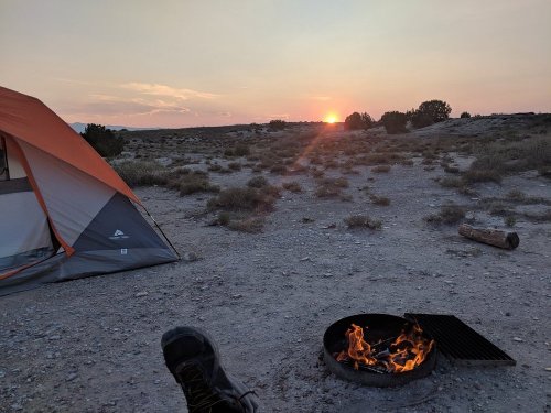 A campsite at sunset, featuring a tent, a campfire, and a vast, open landscape with sparse vegetation.