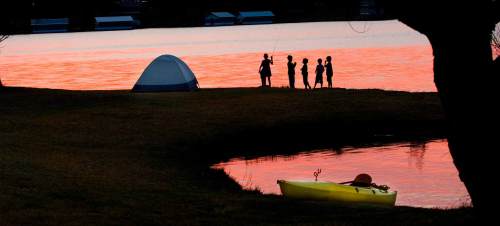 Silhouettes of people by a lake at sunset, with a tent and a kayak in the foreground.