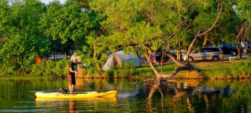 A person stands on a yellow kayak in a calm lake, surrounded by trees and a campsite in the background.