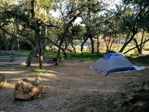 A campsite with a tent set up among trees, a picnic table nearby, and a gravel ground.