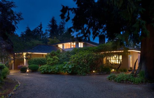 A cozy home illuminated at dusk, surrounded by lush greenery and a gravel driveway.