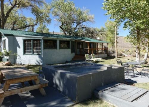 A cozy outdoor area featuring a house, picnic table, and a small hot tub surrounded by trees and a clear blue sky.