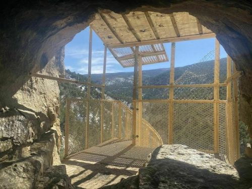 View from inside a rocky cave, showcasing a wooden platform and spiral staircase with a scenic mountain backdrop.