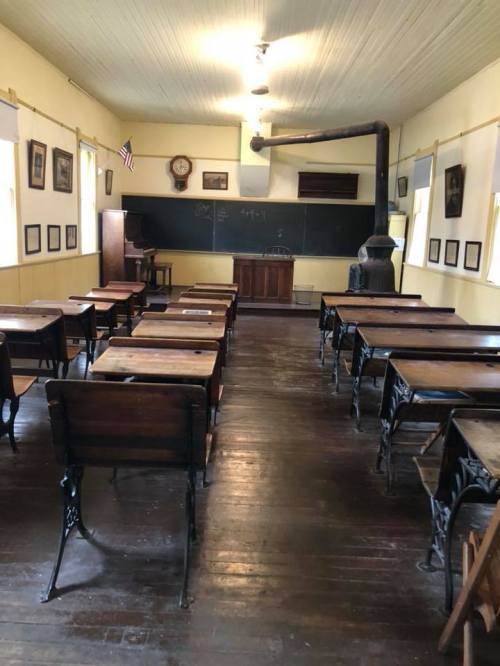 A vintage classroom with wooden desks, a chalkboard, and a potbelly stove, featuring yellow walls and historical decor.