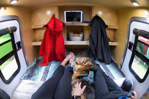 A cozy camper interior with a person’s feet and a dog resting on a bed, surrounded by hanging jackets and shelves.