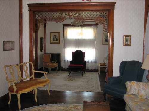 A vintage living room with ornate woodwork, a red armchair, and various seating options, featuring a patterned rug.