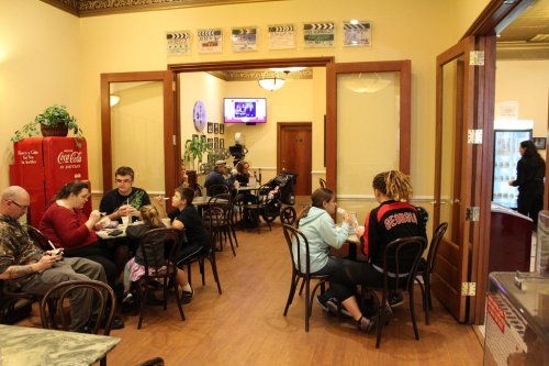 A cozy café interior with families dining at tables, a vintage Coca-Cola fridge, and a TV in the background.