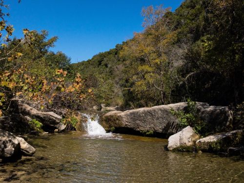 A serene river flows between large rocks, surrounded by trees and a clear blue sky.