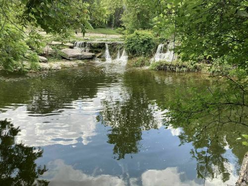 A serene pond with gentle waterfalls, surrounded by lush greenery and reflecting clouds in the water.