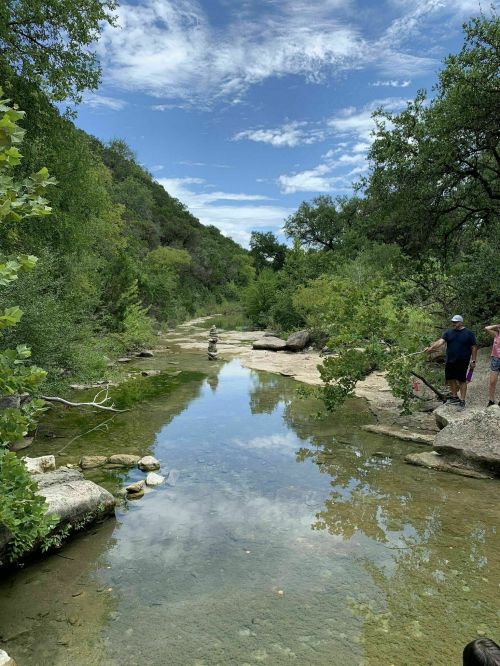 A serene river scene with lush greenery, rocks, and a clear sky reflected in the water, featuring two people by the shore.