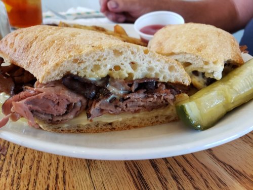 A close-up of a sandwich filled with roast beef, cheese, and mushrooms, served with fries and a pickle on the side.