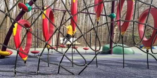 A playground with colorful climbing structures and a child playing in the background, surrounded by trees.
