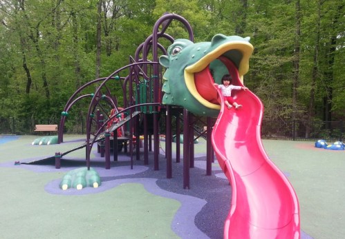 A child slides down a bright red slide shaped like a dragon's mouth at a colorful playground surrounded by trees.