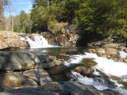 A serene waterfall cascades over rocks into a clear pool, surrounded by lush greenery and trees under a blue sky.