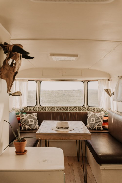 Cozy interior of a vintage camper with a wooden table, cushions, and a view of the outdoors through large windows.