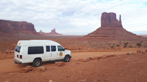 A white van parked on a dirt road with Monument Valley's iconic rock formations in the background under a cloudy sky.